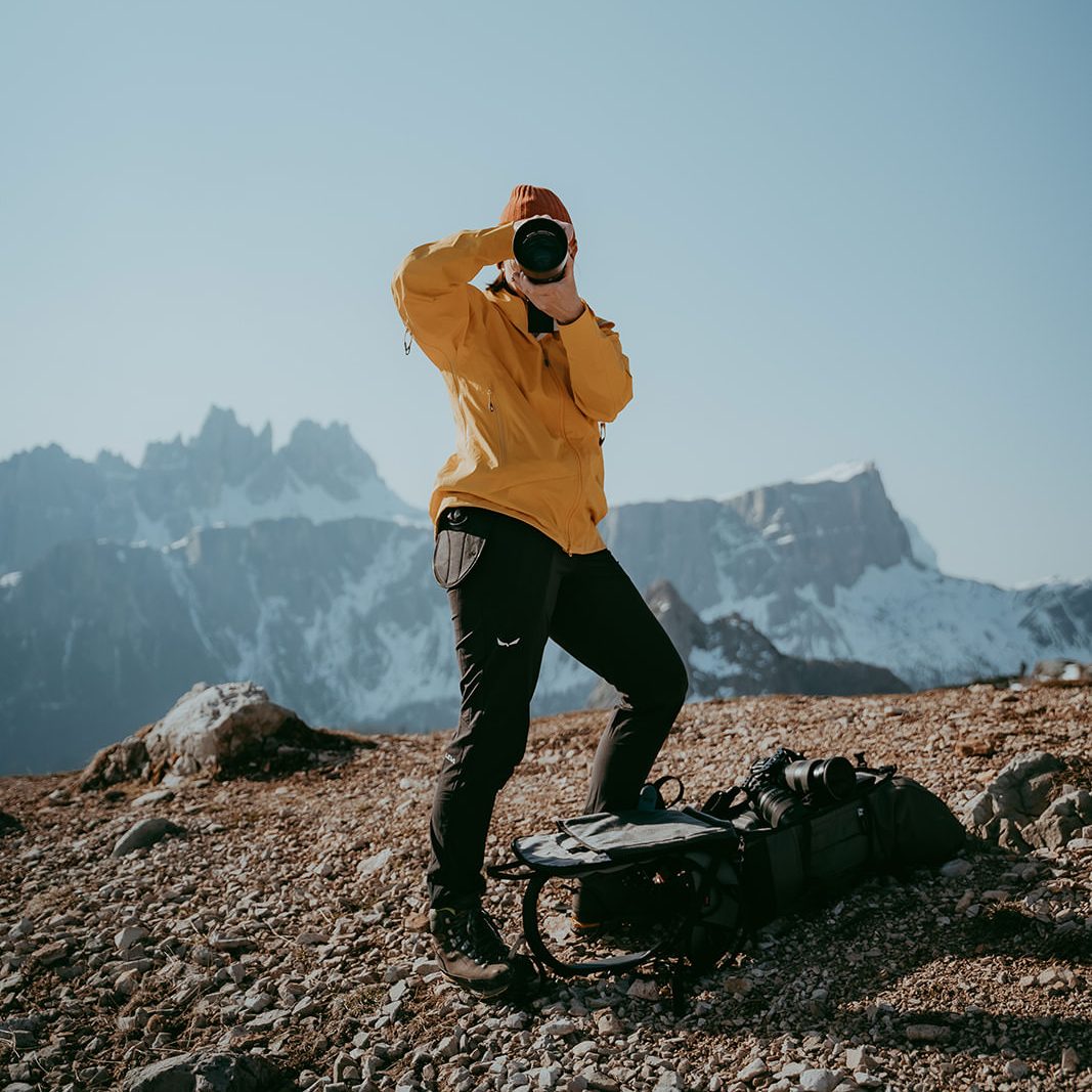 Fotografin Alexandra in gelber Jacke auf Berggipfel mit Kamera, beeindruckende Alpenlandschaft im Hintergrund.