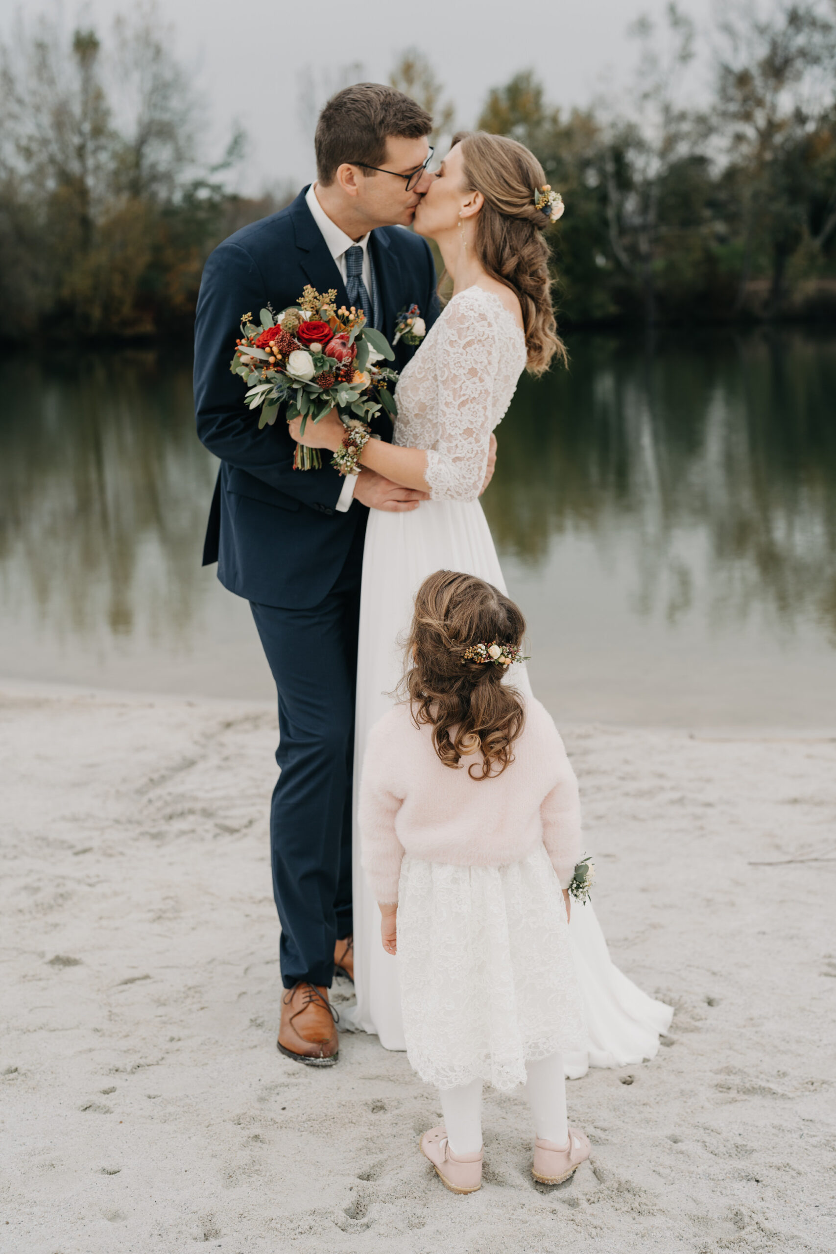 Brautpaar küsst sich bei Hochzeit am Fluss mit Kind im Vordergrund.
