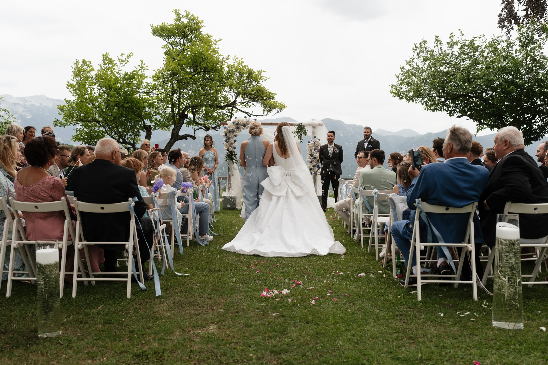 Hochzeit im Freien mit Brautpaar, Gästen und Bergpanorama im Hintergrund.